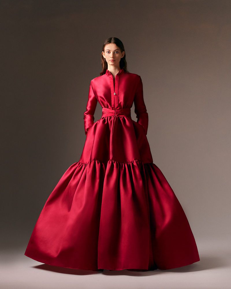 A woman poses in a full-length shot, wearing a long-sleeved, floor-length red gown with a collared top and gathered, ruffled skirt.