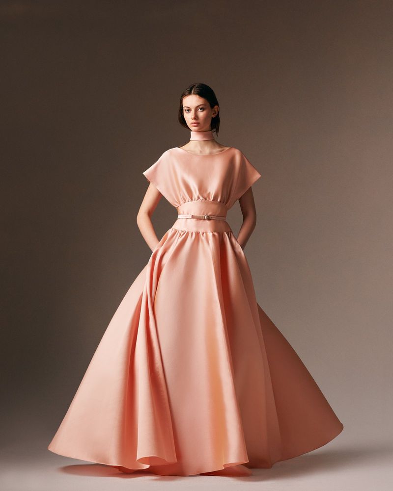 Elegant woman in a peach couture gown with a wide skirt poses in a photo studio with hands in pockets. Fashion editorial.