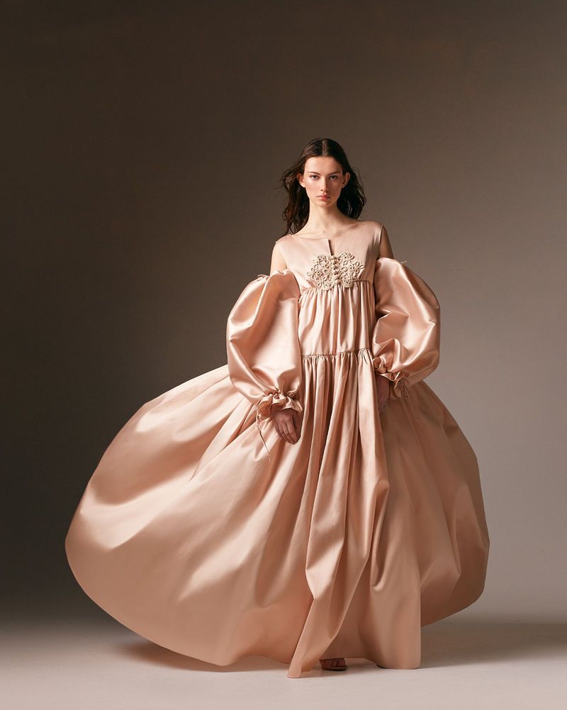 A woman in a long, flowing, light-pink gown with voluminous sleeves stands in a neutral studio. Fashion photography, studio lighting.