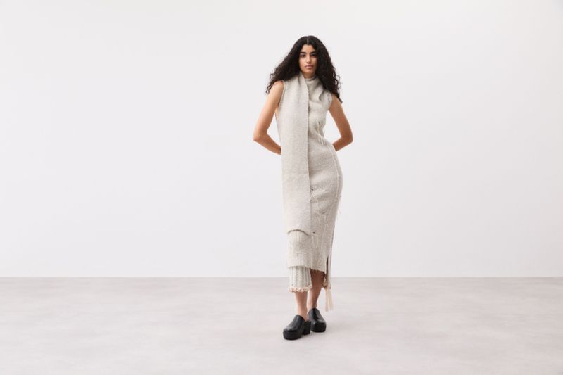 Adult woman in a knit dress and scarf poses in a white studio, standing in black platform shoes with her hands on her hips.