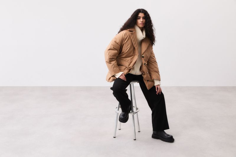 Fashionable woman in a brown puffer jacket and black trousers sitting on a silver stool in a bright, minimalist studio setting.