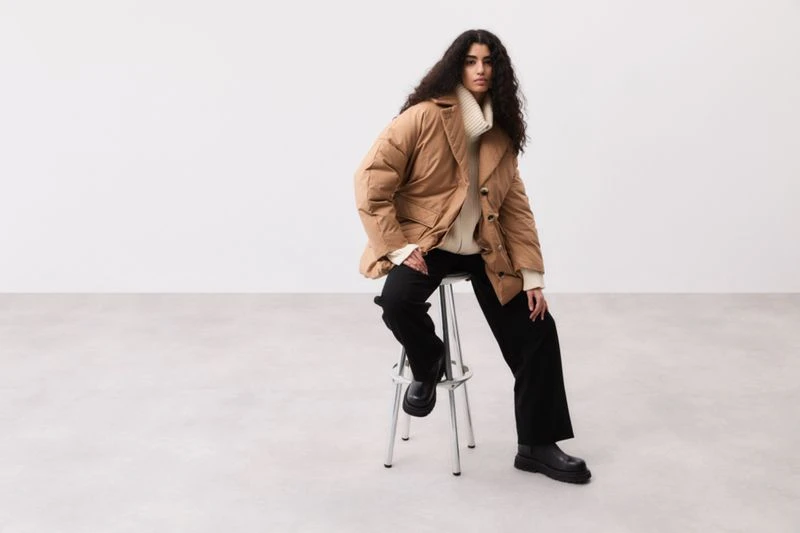 Fashionable woman in a brown puffer jacket and black trousers sitting on a silver stool in a bright, minimalist studio setting.