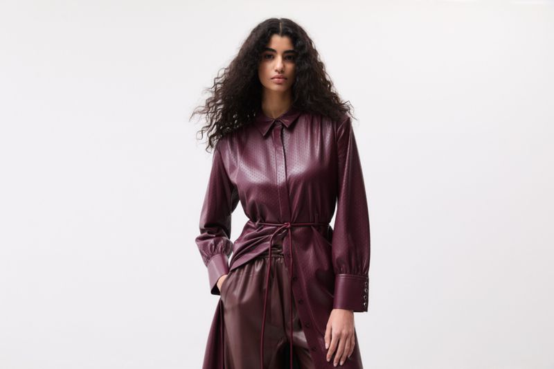 A stylish adult female wearing a maroon faux leather dress and pants posing in a photo studio with a soft lighting background.