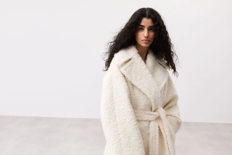 A woman with curly hair wears an off-white wrap coat with a tie belt against a plain white backdrop in a fashion studio shot.