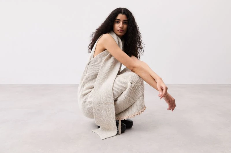 A woman with dark curly hair poses crouched down in a photo studio wearing a light beige knitted vest and pants outfit. Indoor fashion shot.