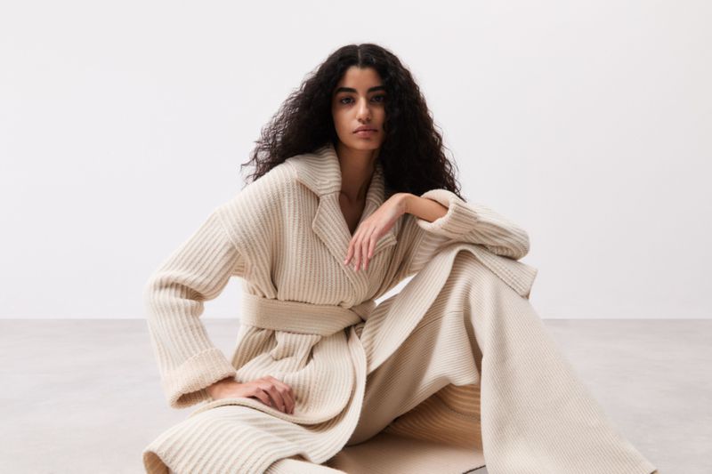A woman with dark curly hair poses indoors wearing a cream-colored, belted, ribbed cardigan and matching pants in a fashion shot.