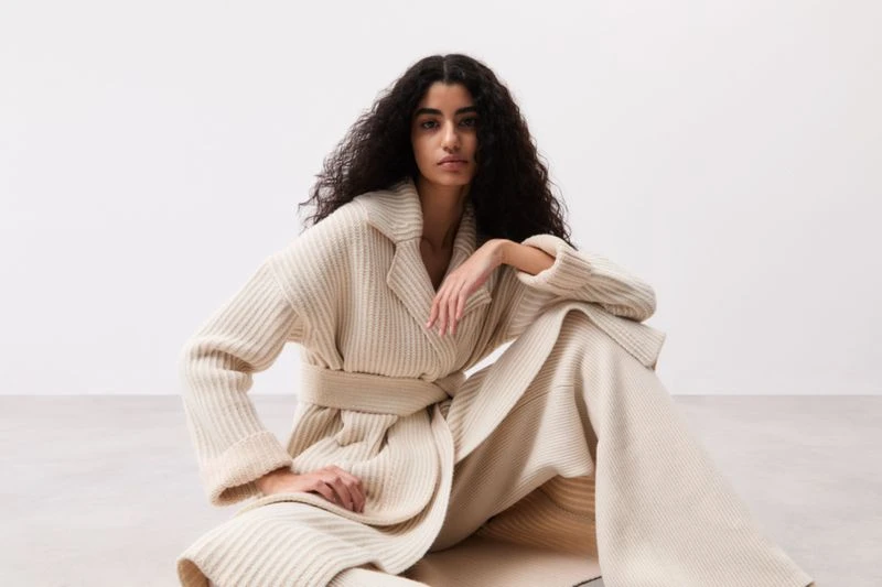 A woman with dark curly hair poses indoors wearing a cream-colored, belted, ribbed cardigan and matching pants in a fashion shot.