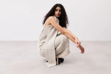 Model Sephora Mosbah crouching in a beige knit sleeveless outfit with a long draped scarf in a minimalist studio setting.