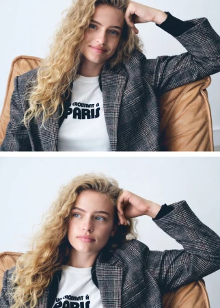 Blonde woman with curly hair poses in an armchair wearing a plaid jacket and 'Croissant a Paris' t-shirt in a portrait shot.