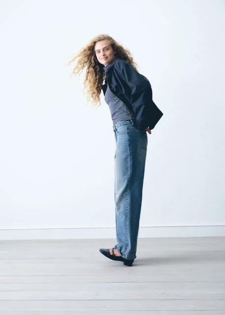 Full-length shot of a smiling woman with curly hair, wearing a striped shirt, jacket, blue jeans, and flats, posing against a white background.