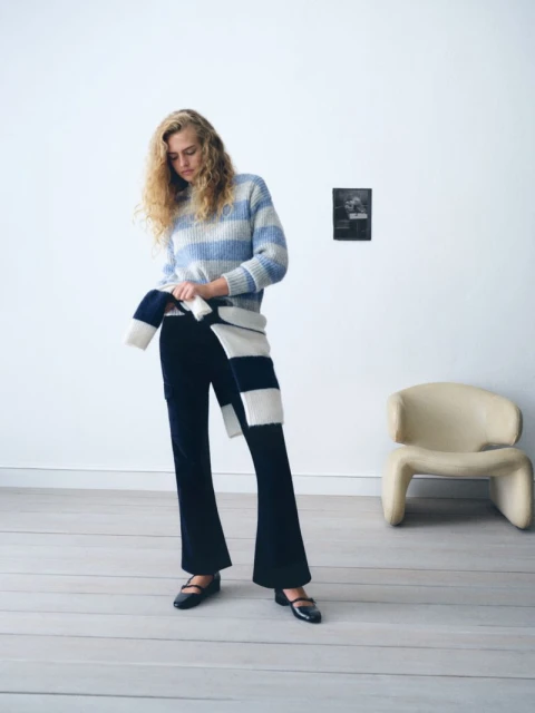 A woman poses indoors wearing a blue and white striped sweater and black pants, with a striped scarf, in a fashion editorial setting.
