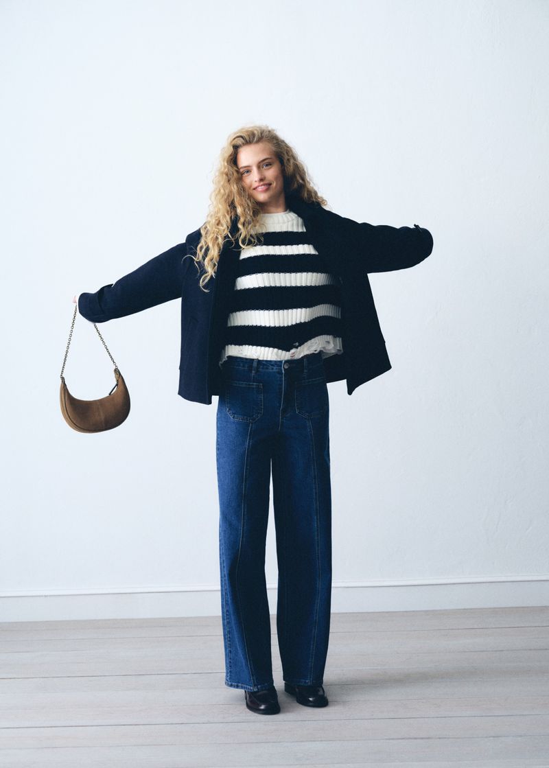 A blonde woman in jeans, a striped sweater, and a jacket poses with a brown bag against a white background in a studio setting.