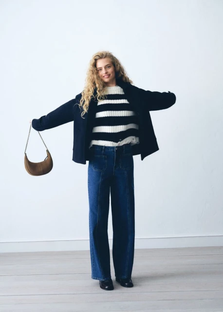 A blonde woman in jeans, a striped sweater, and a jacket poses with a brown bag against a white background in a studio setting.