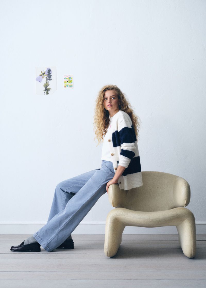 A fair-skinned woman with curly blonde hair sits on a tan chair in a bright photo studio wearing wide leg jeans and a striped cardigan.