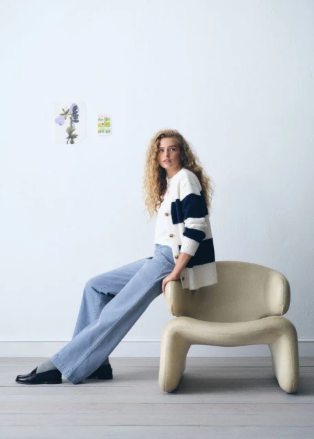 A fair-skinned woman with curly blonde hair sits on a tan chair in a bright photo studio wearing wide leg jeans and a striped cardigan.