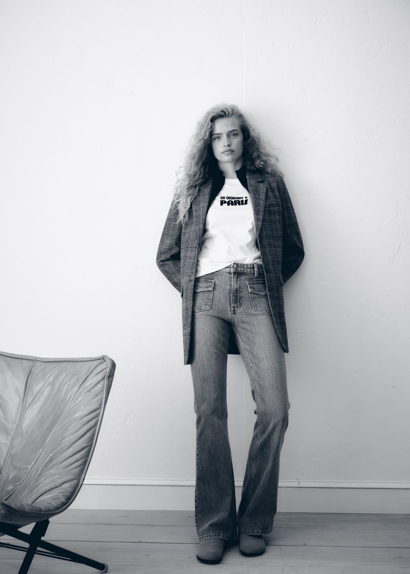 A full-length black-and-white portrait shows a slender woman with wavy hair wearing jeans, a t-shirt, and a blazer leaning against a white wall.