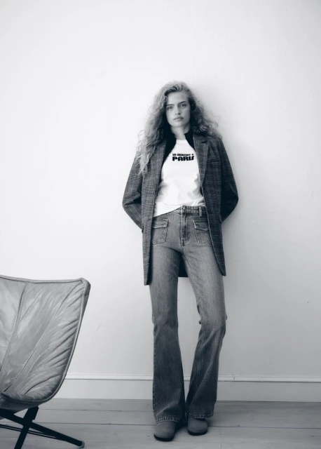 A full-length black-and-white portrait shows a slender woman with wavy hair wearing jeans, a t-shirt, and a blazer leaning against a white wall.
