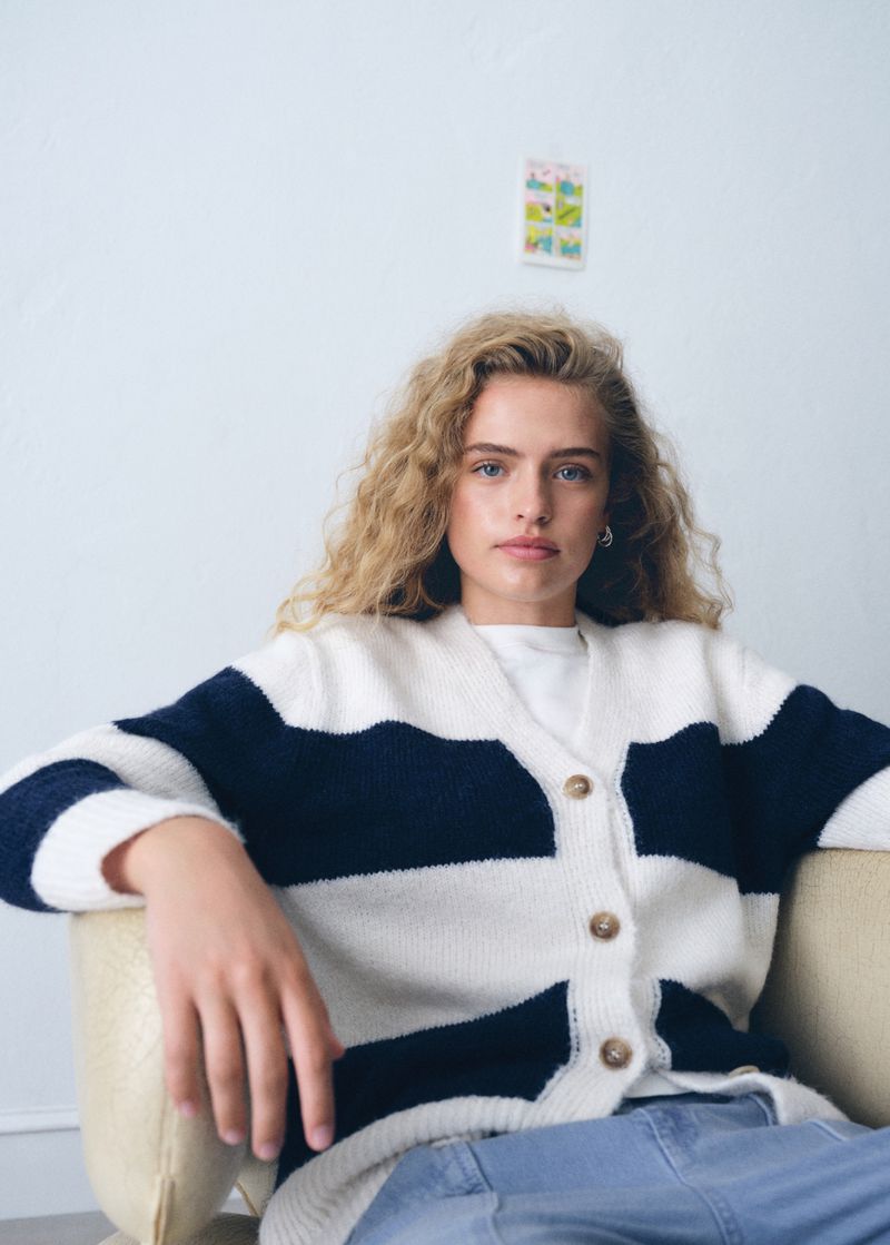 Young female model with curly blonde hair seated in an armchair wearing a striped cardigan and blue denim jeans.