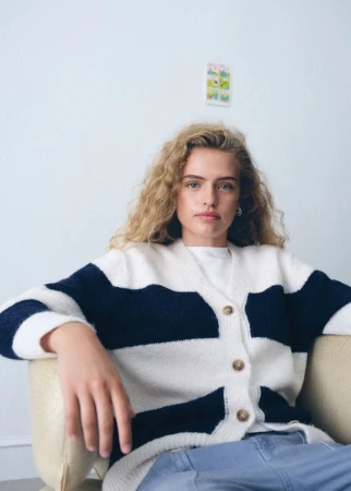 Young female model with curly blonde hair seated in an armchair wearing a striped cardigan and blue denim jeans.