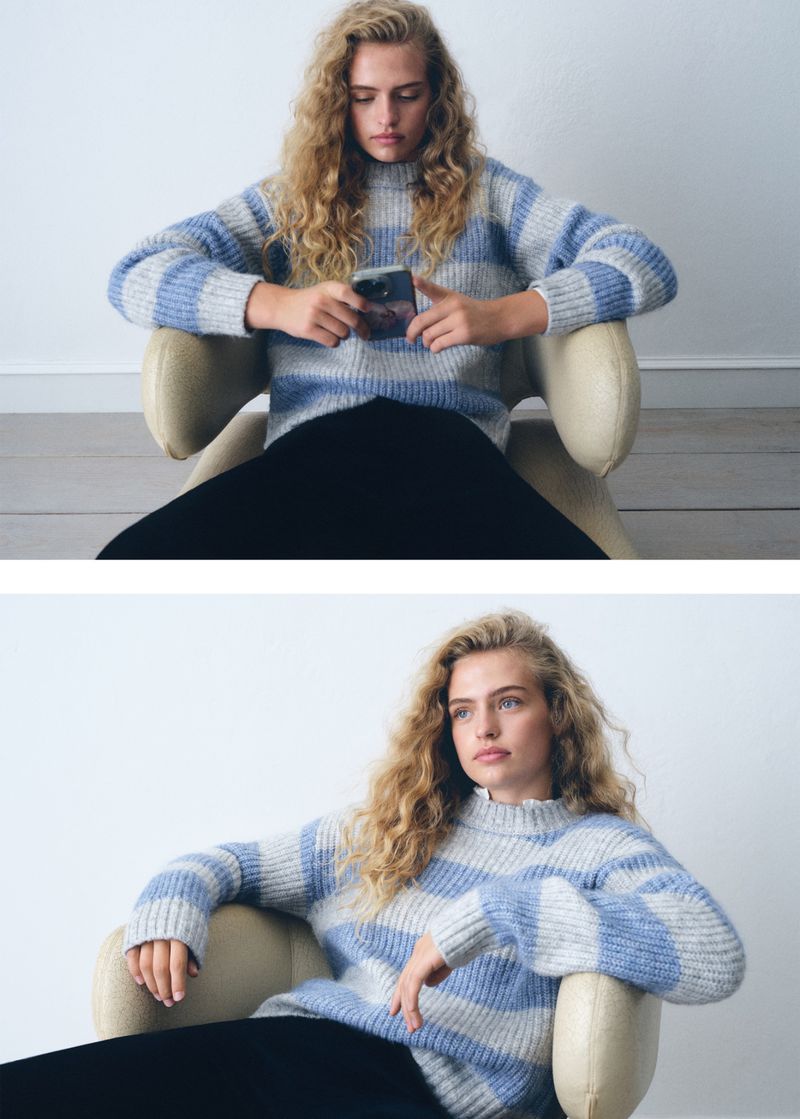 A woman with curly blonde hair sits in a chair, wearing a striped sweater and looking at a smartphone, captured in a studio setting.