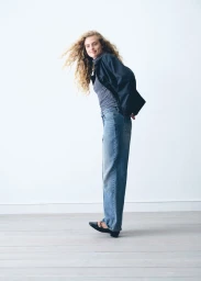 Model with long curly hair wearing a dark jacket, striped top, and blue jeans against a white background.