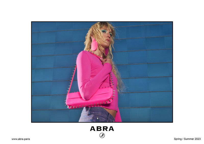Fashion shot of a blonde adult woman in a hot pink top with matching spiked handbag against a blue geometric wall in daylight.
