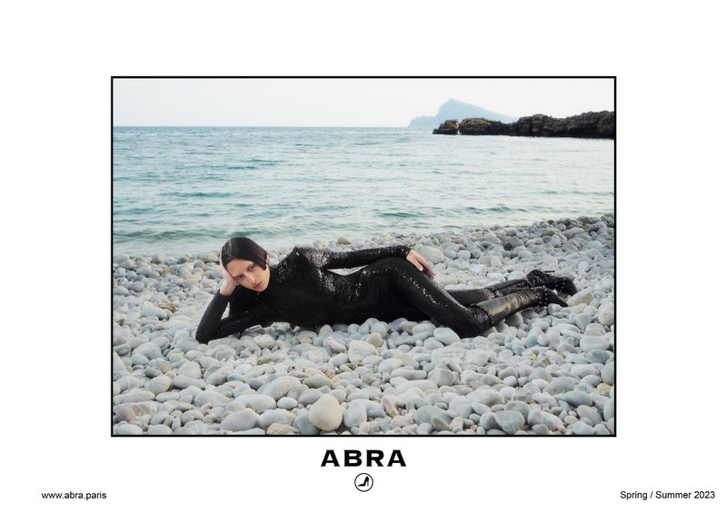 A woman in a black catsuit poses on a rocky beach by the ocean. She is lying down facing left with a mountain in the background.