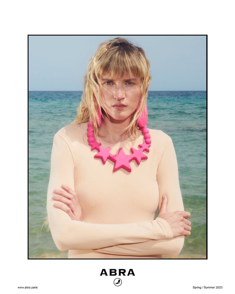 Adult woman with blonde hair poses in front of ocean wearing a pink star necklace and pink feather earrings in an outdoor setting.