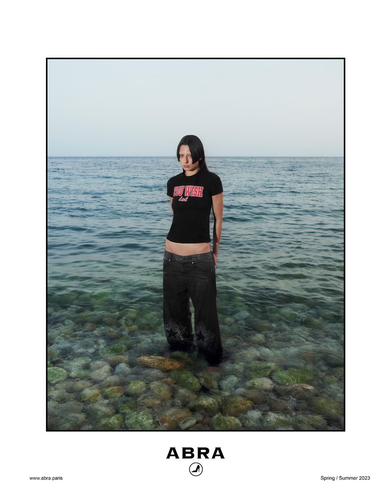 A woman in a black t-shirt and loose dark pants stands in shallow water by the sea, creating a serene fashion-editorial image.