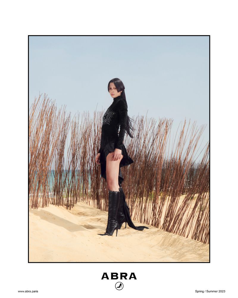 Fashion shot of a woman in a black dress and boots posing on a sandy beach with tall grass, under a clear sky, promoting apparel.