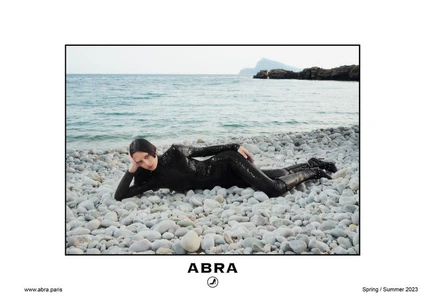 A model in a shimmering black catsuit reclines on a stony pebble beach with the calm turquoise ocean in the background.
