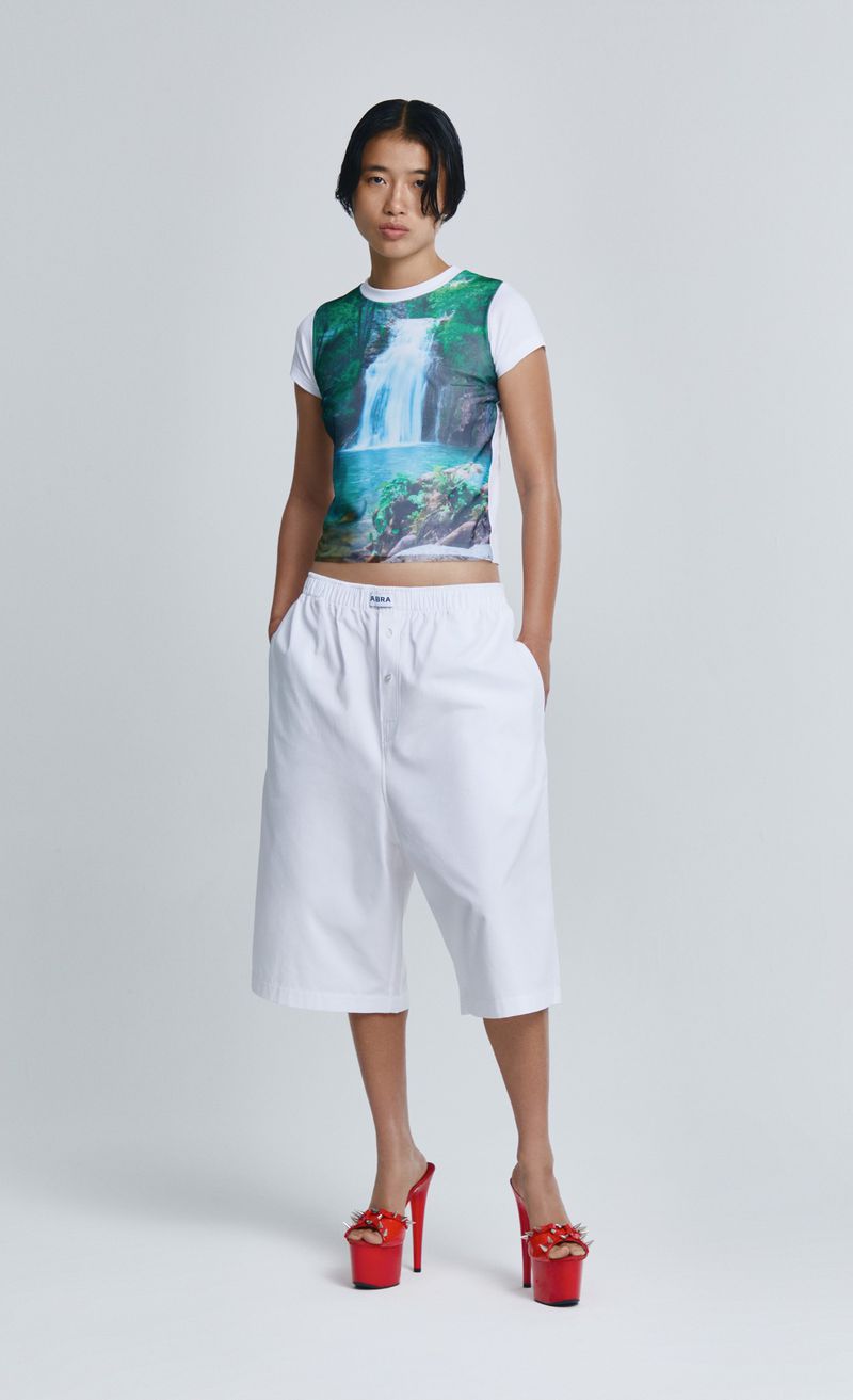An Asian woman stands in a studio wearing a waterfall-print t-shirt, wide white shorts, and platform red shoes against a white background.