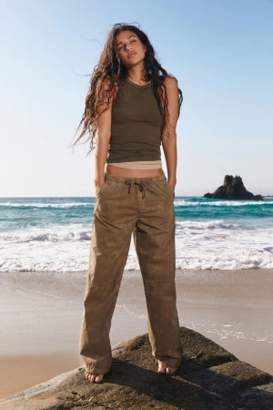 Model Tamar David posing on a coastal rock in an olive tank top and loose khaki trousers for the Pull & Bear S/S 24 lookbook.
