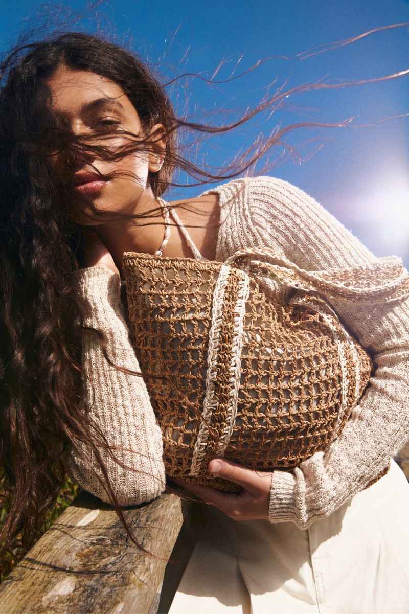 Woman holds a brown straw bag on a sunny day. The woman has dark curly hair, a light complexion, and wears a light beige sweater.