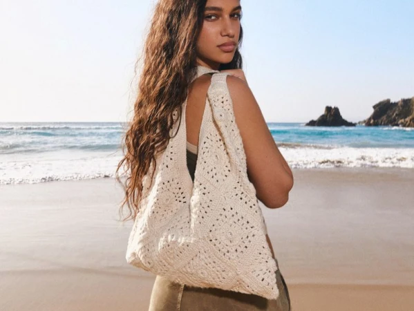Woman on a beach, carrying a crochet tote bag over her shoulder, with the ocean and sky in the background on a sunny day.