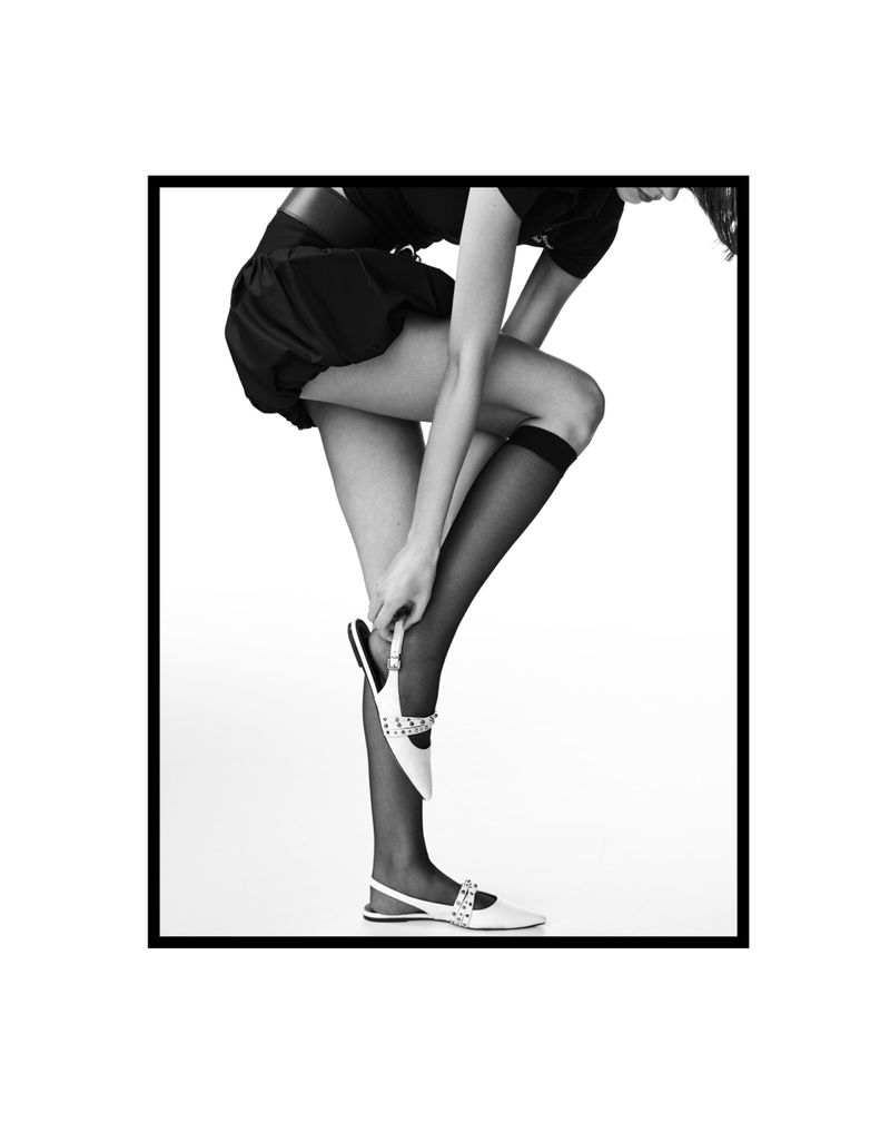 A slim woman in a black dress bends over while putting on a jeweled white shoe in a black and white studio fashion shot.