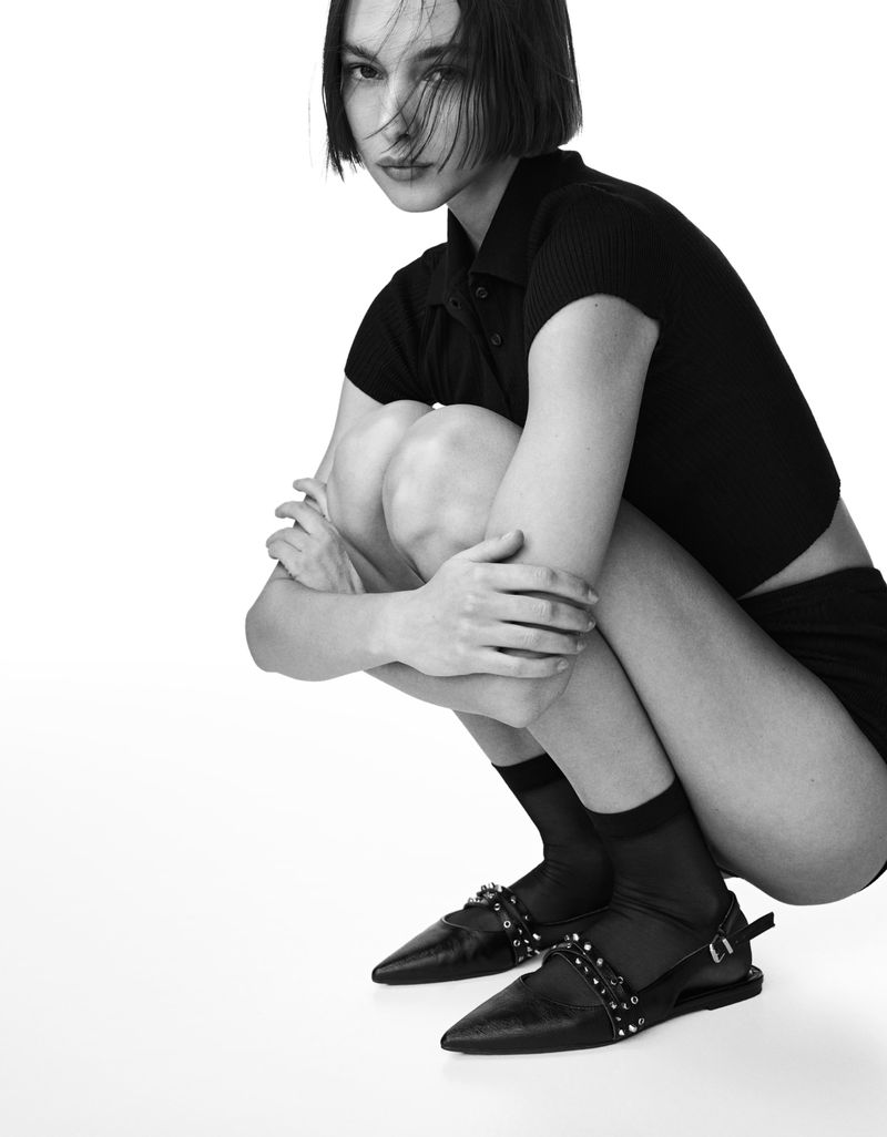 Monochromatic studio shot of a woman crouching in a black crop top, shorts, socks, and embellished flats against a white background.