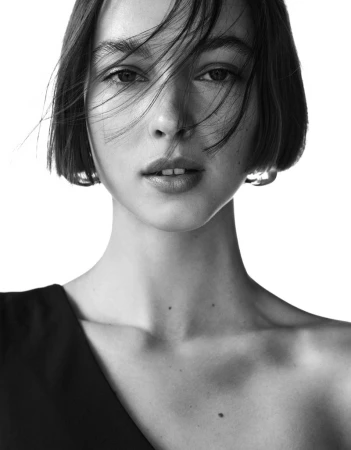 Black and white close-up portrait of model Alida Aciu with a short bob haircut and a bare shoulder against a white background.