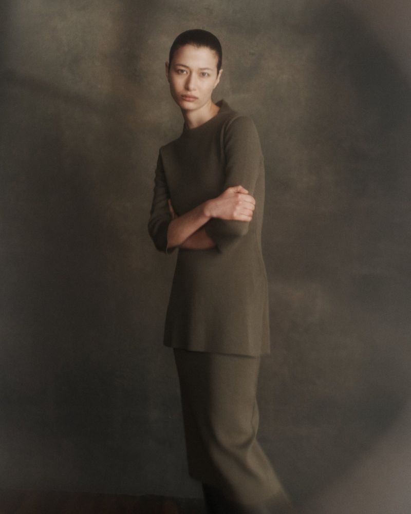 Portrait of a woman in a minimalist green knit suit with arms crossed, posing in a dimly lit studio with a blurred background.