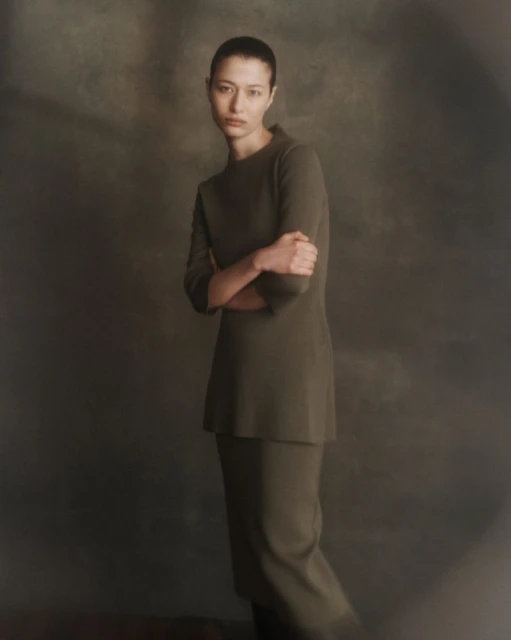 Portrait of a woman in a minimalist green knit suit with arms crossed, posing in a dimly lit studio with a blurred background.