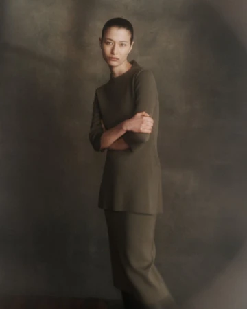 Portrait of a woman in a minimalist green knit suit with arms crossed, posing in a dimly lit studio with a blurred background.