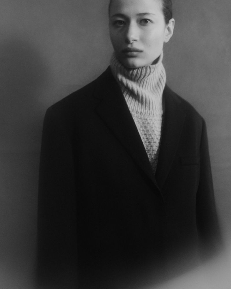 Black and white portrait of an Asian woman in a black coat and tan turtleneck sweater, with a grey studio background.