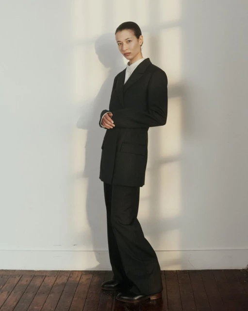 Fashionable adult female posing in a black pantsuit, standing in front of a textured white wall with soft, diffused lighting and shadows.