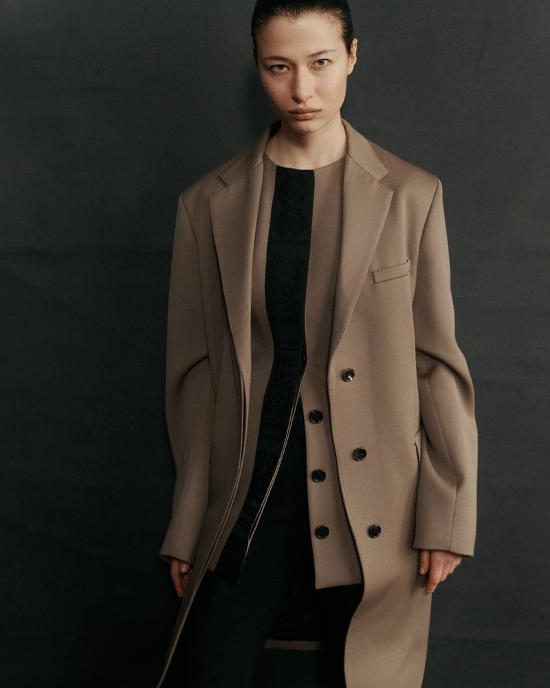 A young woman with slicked-back hair poses in a studio wearing a tan coat and black dress, her gaze fixed directly at the camera.