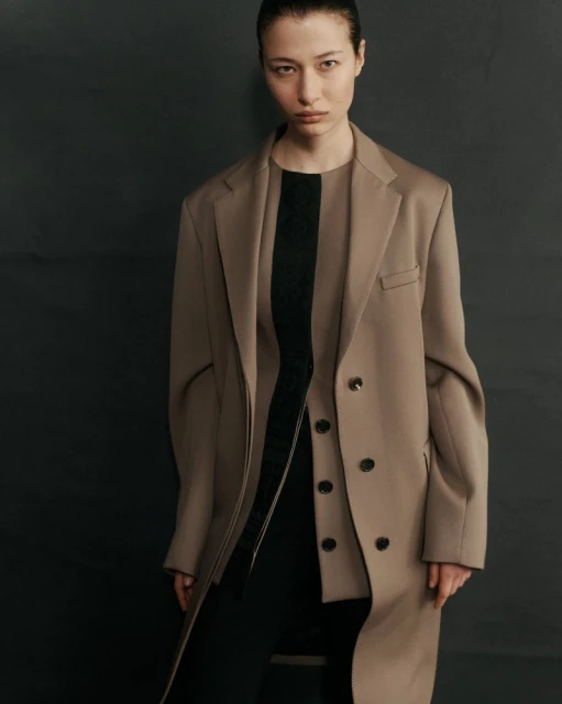A young woman with slicked-back hair poses in a studio wearing a tan coat and black dress, her gaze fixed directly at the camera.