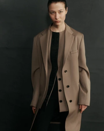 A young woman with slicked-back hair poses in a studio wearing a tan coat and black dress, her gaze fixed directly at the camera.