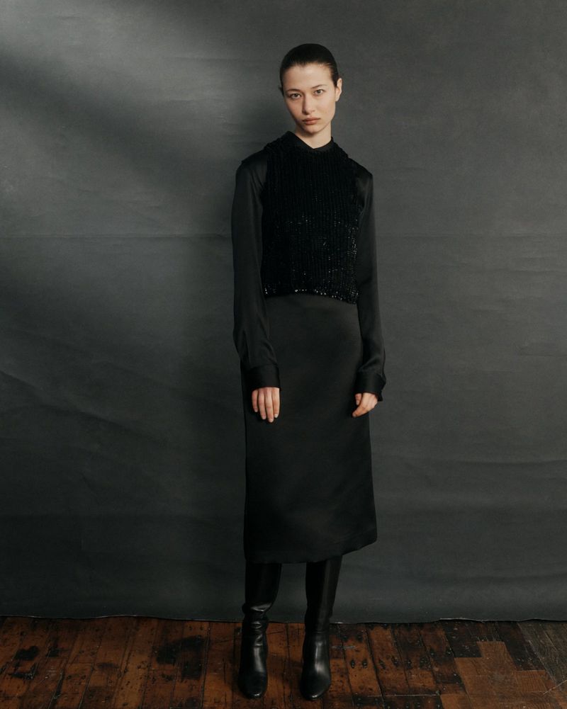 Model Leyla Greiss wearing a black satin midi dress, textured knit vest, and knee-high leather boots against a dark grey backdrop.