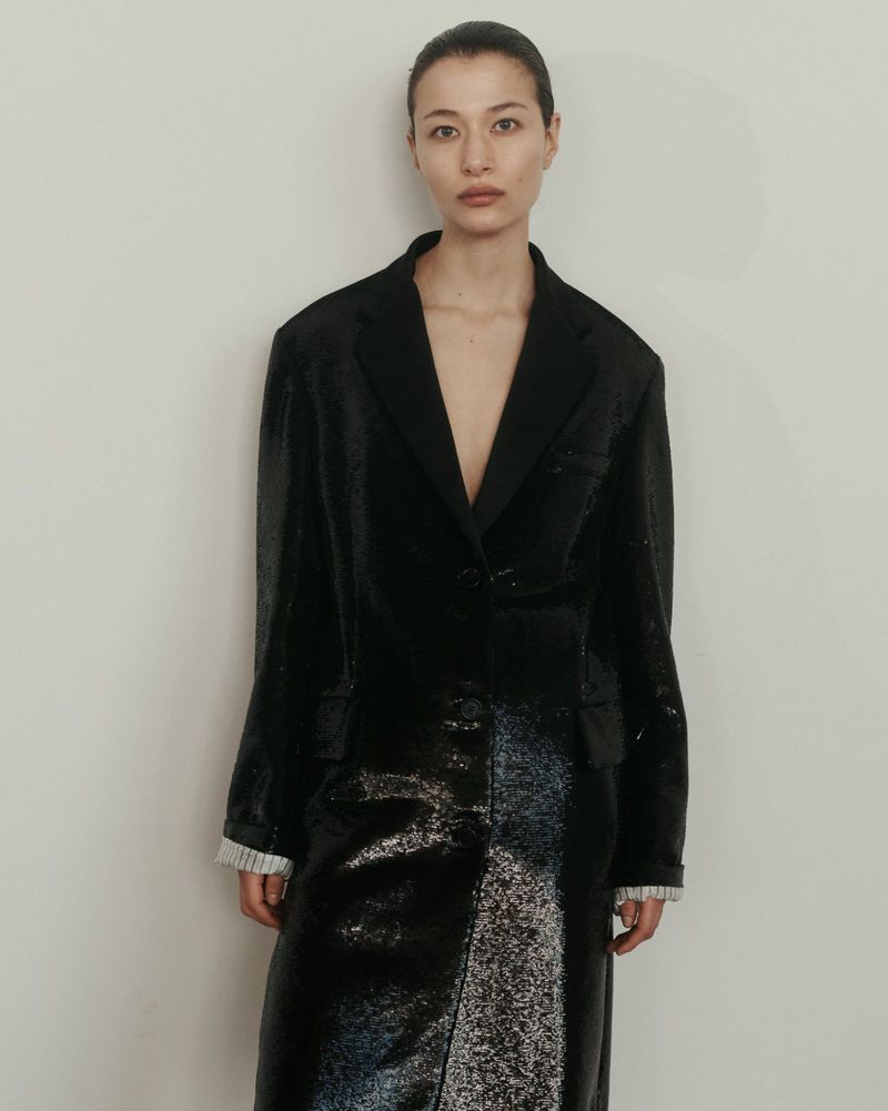 An Asian woman model poses in a long black shiny coat against a neutral wall, fashion-forward and elegant in a studio setting.