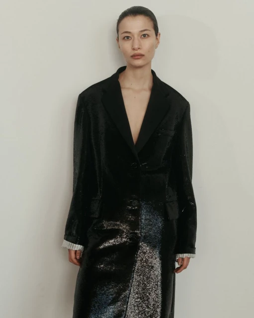 An Asian woman model poses in a long black shiny coat against a neutral wall, fashion-forward and elegant in a studio setting.