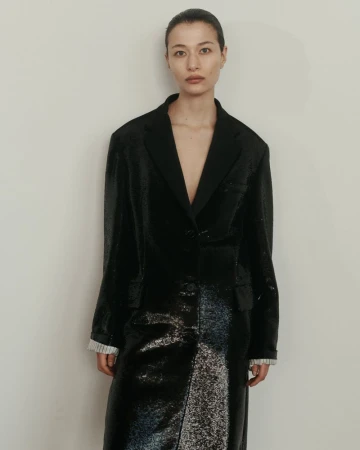 An Asian woman model poses in a long black shiny coat against a neutral wall, fashion-forward and elegant in a studio setting.